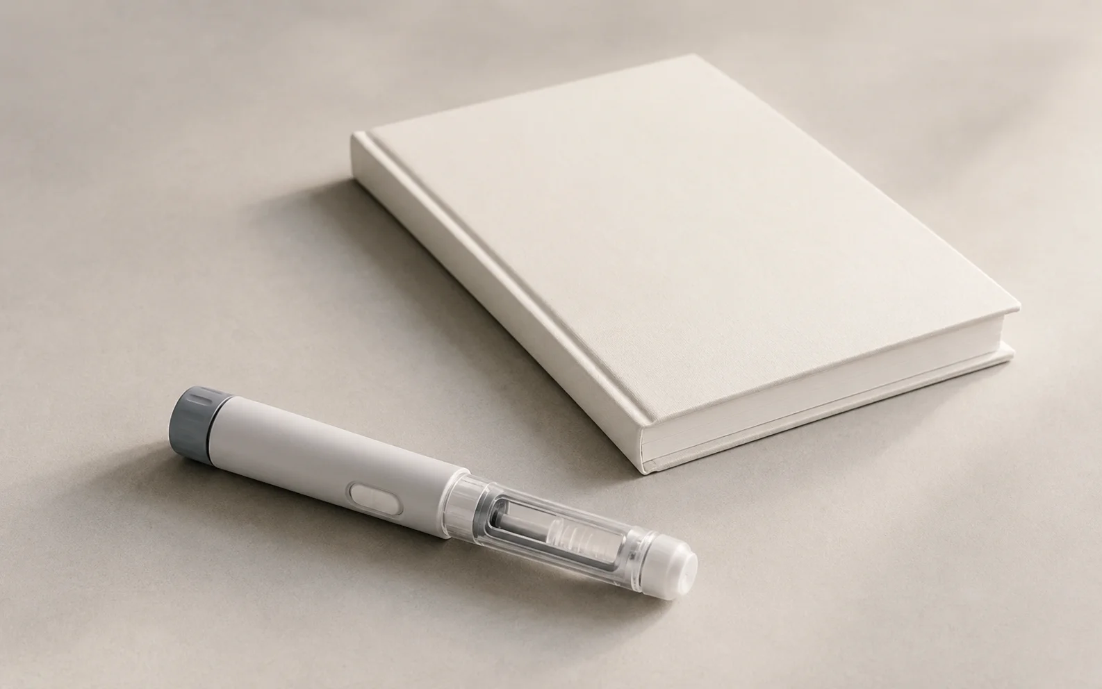 A plain unbranded medical injector pen beside a blank notebook on a calm neutral surface